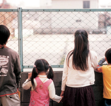 Four children facing away from the camera, looking through a chainlink fence