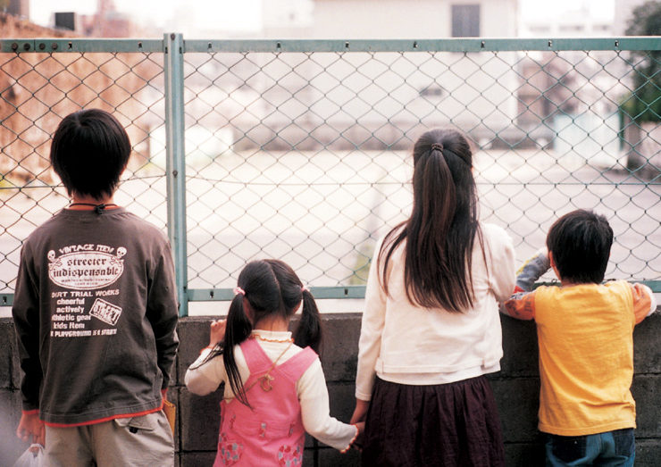 Four children facing away from the camera, looking through a chainlink fence