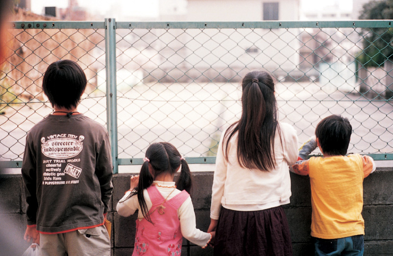 Four children facing away from the camera, looking through a chainlink fence