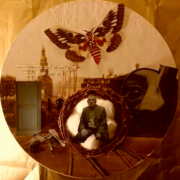 Collage with a photo of an old man in the center, encircled by a nest of twigs, a cityscape is behind him and there is a large moth at the top