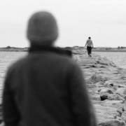 Black and white photo of a person on a breakwater wall looking out at another person in the distance