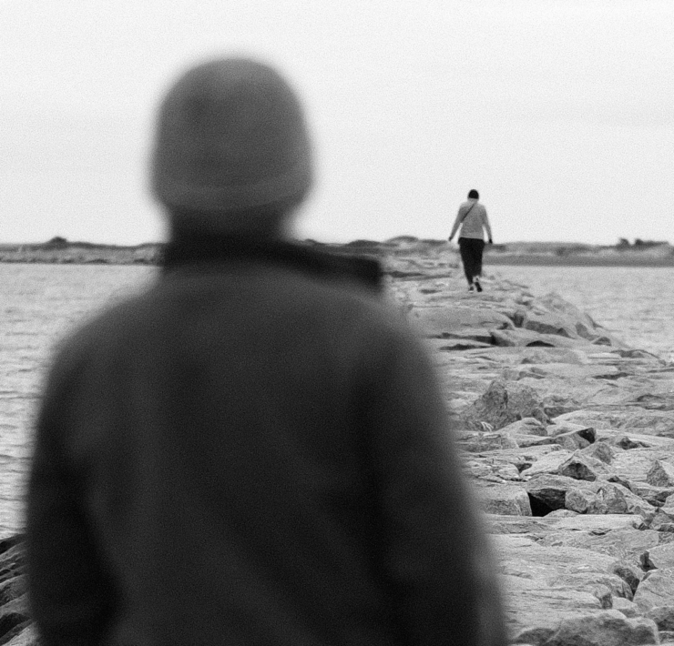 Black and white photo of a person on a breakwater wall looking out at another person in the distance