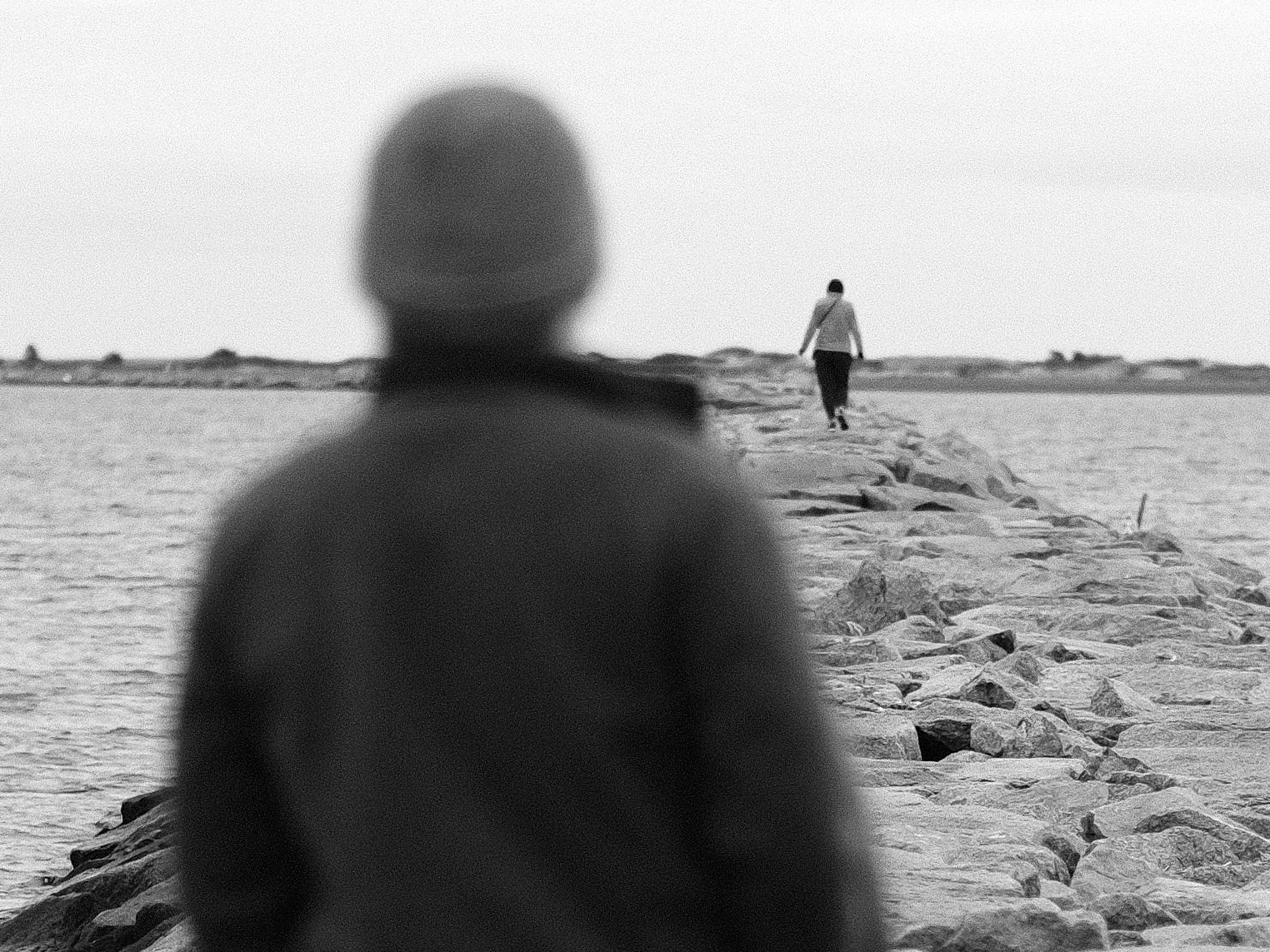 Black and white photo of a person on a breakwater wall looking out at another person in the distance