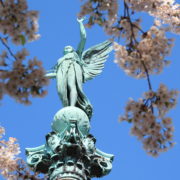 Photo of a green angel statue as seen from below pink cherry blossom branches