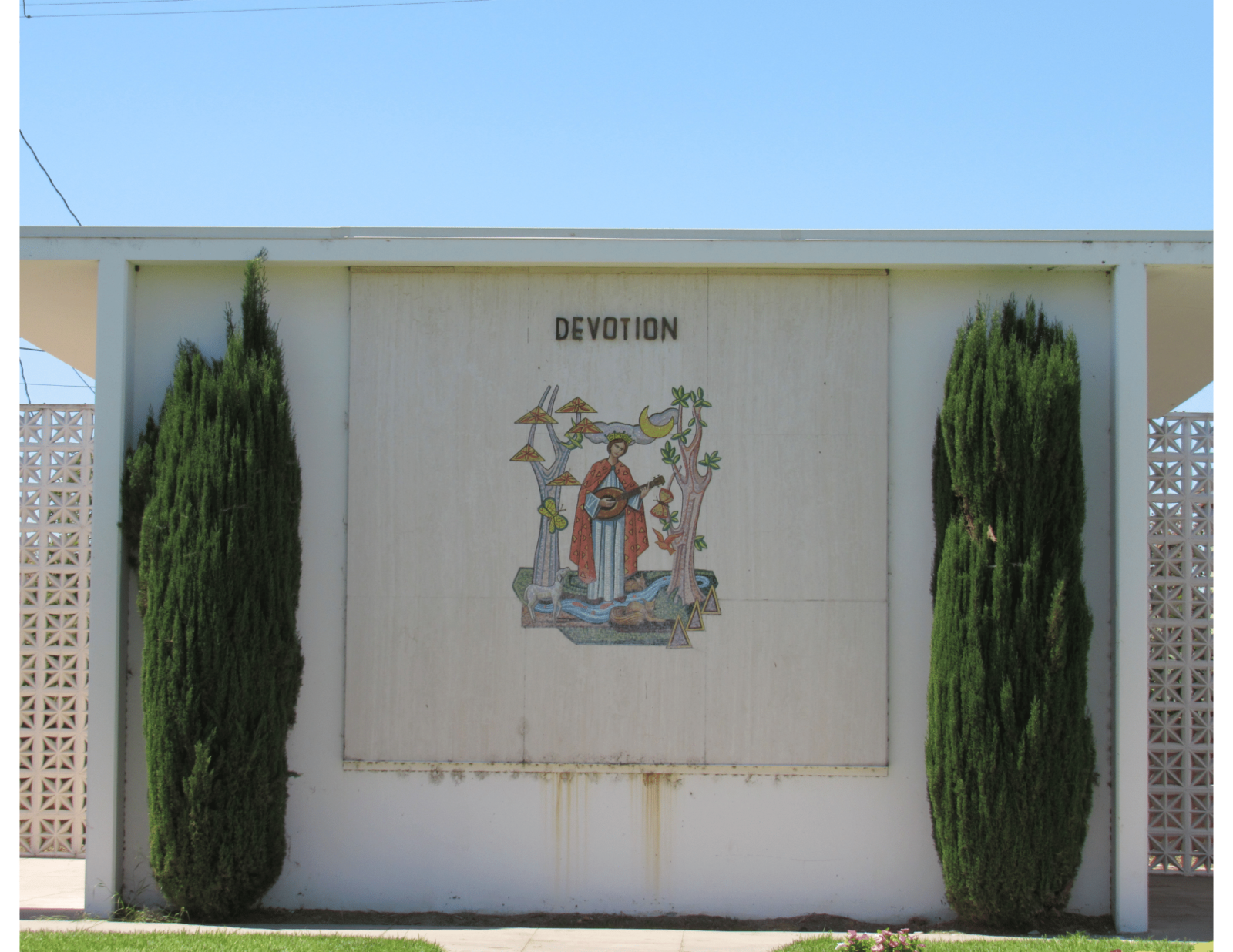 Photo of the side of a building flanked by two juniper trees, in the center of the wall is a depiction of a saint with the word Devotion above