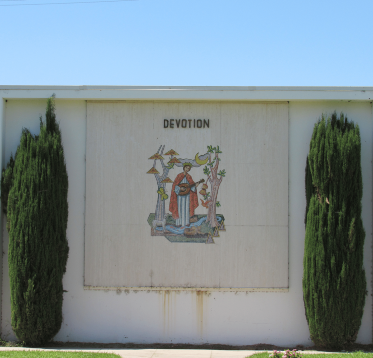 Photo of the side of a building flanked by two juniper trees, in the center of the wall is a depiction of a saint with the word Devotion above